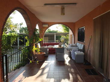 The image depicts a covered outdoor patio with arched brick walls, featuring a couch, table, and potted plants, bathed in sunlight.