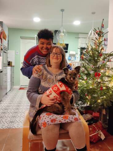 The image depicts a woman sitting on a chair, cradling a small dog adorned with a festive holiday sweater, surrounded by a Christmas tree and presents in a cozy home setting.