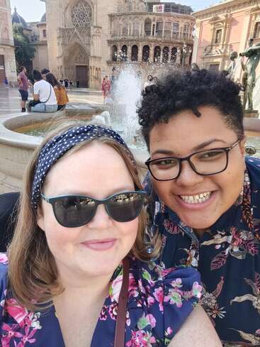 The image shows two people posing for a selfie in front of a fountain and historic buildings, with a church and statue visible in the background.