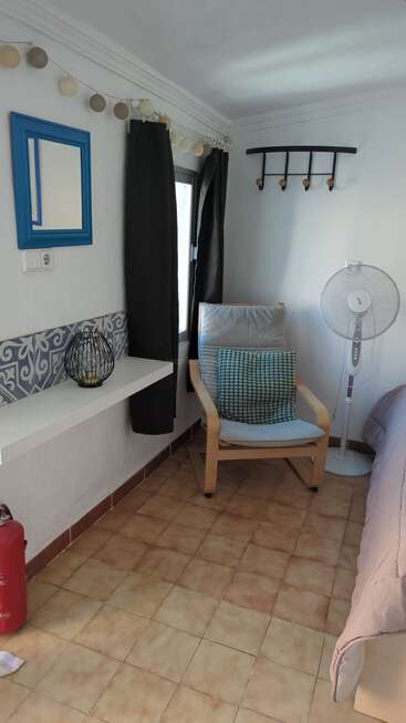 Un coin douillet composé d'une chaise en bois avec des coussins, d'un ventilateur sur pied, de crochets muraux, d'un miroir à cadre bleu, de guirlandes lumineuses, de bougies décoratives et d'un sol carrelé.