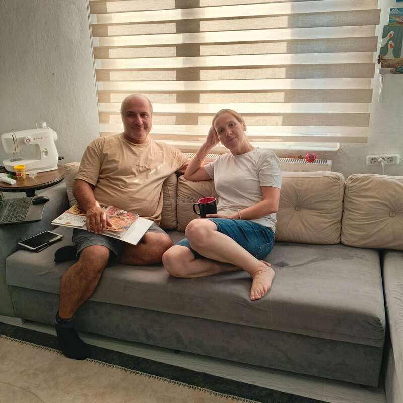 A man and woman sit comfortably on a gray couch; she holds a mug, he holds a magazine. There’s a sewing machine, laptop, and phone nearby.