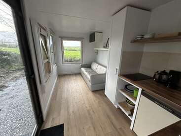 This image shows a tiny house interior with wooden flooring, a small kitchen area, open shelving, a cozy sofa, large windows, and a beautiful green outdoor view.