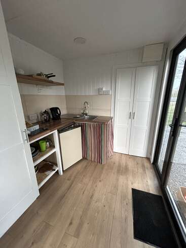 This is a small, cozy kitchen with wooden floors, open shelves, a striped curtain under the sink, minimal appliances, white cabinets, and a glass door.