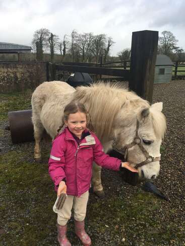A smiling young girl in a pink jacket and boots stands beside a white pony, gently brushing it, outdoors on a cloudy day at a farm.