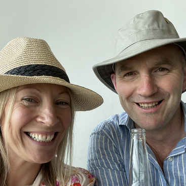A smiling woman and man, both wearing hats, enjoy a cheerful moment together. The man holds a glass bottle, and they sit against a plain background.