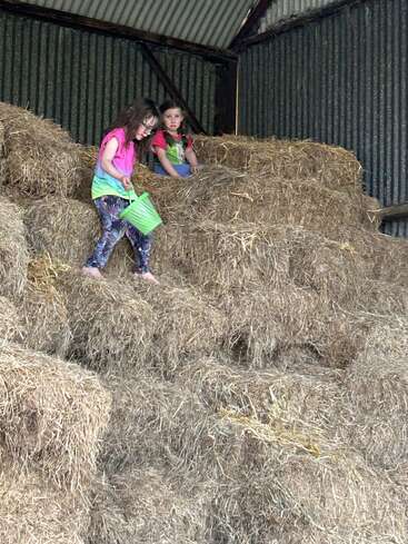 Two young girls are playing on a large stack of hay bales in a barn, one holding a green basket, both dressed in colorful clothes.