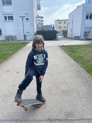 Un jeune enfant, vêtu d'un sweat à capuche et de bottes, se tient debout avec assurance sur un skateboard, sur un chemin pavé entre deux immeubles, profitant d'un moment en plein air.