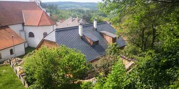 A imagem mostra uma área residencial tranquila com várias casas com telhados e chaminés escuros, em meio a uma vegetação exuberante e colinas ao fundo.