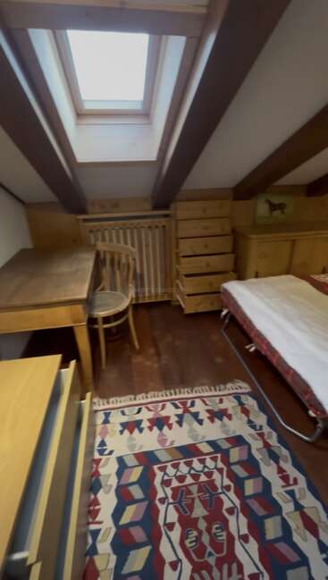 Cette chambre mansardée confortable dispose d'une lucarne, de poutres en bois apparentes, d'un bureau en bois avec une chaise, d'un tapis à motifs, d'un lit et de plusieurs commodes.