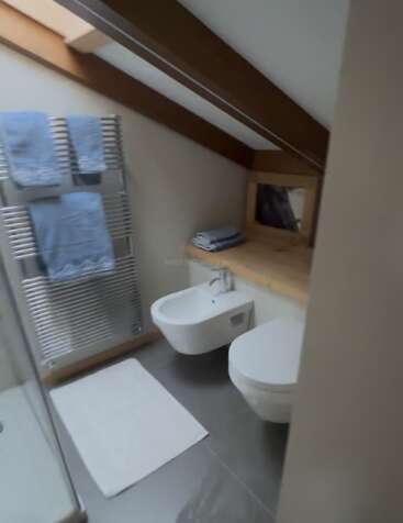 Cette salle de bains présente un plafond incliné avec des poutres en bois, un porte-serviettes avec des serviettes bleues, un bidet, des toilettes, un petit miroir, un comptoir, une douche et un sol carrelé gris.