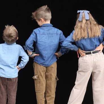 Três crianças estão de costas para a câmera contra um fundo preto, vestindo camisas jeans. A menina tem um laço no cabelo. Mãos nos quadris.