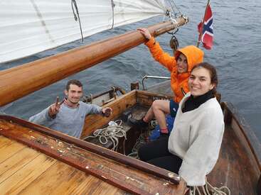 Tres amigos navegan en un barco de madera, sonriendo y disfrutando de su tiempo. Uno de ellos hace el signo de la paz, mientras la bandera noruega ondea de fondo.