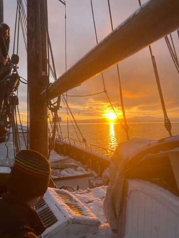 Uma pessoa senta em um barco coberto de neve ao pôr do sol. A luz laranja reflete na água e no barco, com cordas, mastros e uma paisagem tranquila de inverno à vista.
