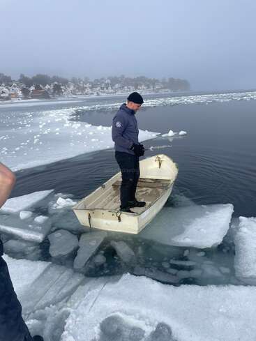 Um cara está em um pequeno barco flutuando em águas geladas, cercado por grossas camadas de gelo. Casas e árvores cobertas de neve são visíveis no fundo nebuloso.
