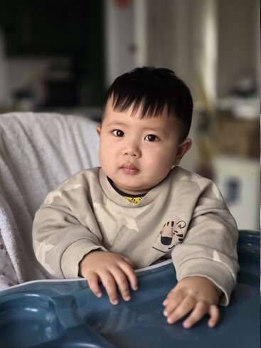 Un petit enfant aux cheveux noirs et raides est assis dans une chaise haute ; il porte un pull beige orné d'un motif de tigre de dessin animé et regarde pensivement l'appareil photo.