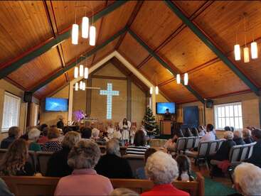 A congregação de uma igreja assiste a uma apresentação de Natal com crianças. Uma árvore decorada, uma cruz e um teto de madeira contribuem para a atmosfera festiva e calorosa.