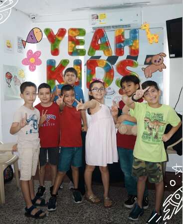 Six enfants joyeux posent devant une enseigne colorée « YEAH KIDS », entourés de dessins muraux ludiques, faisant le signe de la paix et souriant avec enthousiasme.