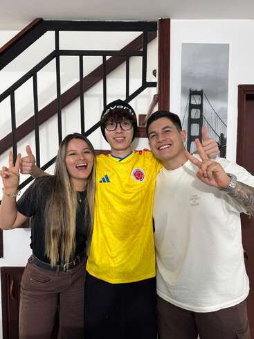 Trois jeunes adultes posent joyeusement, souriants et faisant le signe de la paix. Ils se tiennent à l'intérieur près d'un escalier, l'un d'eux porte un maillot de football colombien, dégageant ensemble une atmosphère joyeuse et amicale.