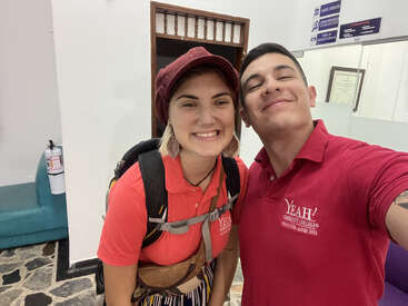 Deux jeunes adultes souriants posent pour un selfie à l'intérieur, tous deux vêtus de t-shirts rouges « YEAH ! ». La femme porte un sac à dos et un chapeau, l'homme arbore un large sourire.