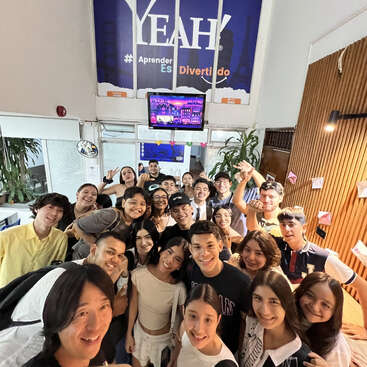 A large group of smiling young people pose for a selfie indoors. Behind them, a sign reads "YEAH! Aprender ES Divertido." The atmosphere appears friendly and cheerful.