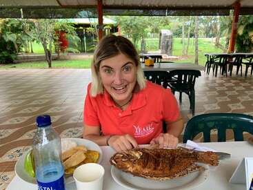 Une femme vêtue d'une chemise rouge est assise, enthousiaste, à la table d'un restaurant en plein air, prête à déguster un poisson frit entier accompagné de riz, de bananes plantains et d'une bouteille d'eau.