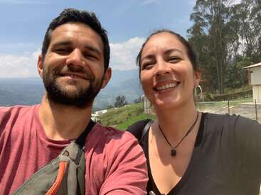 Un homme et une femme souriants prennent un selfie en plein air par une journée ensoleillée, avec des collines verdoyantes, des arbres, un ciel bleu et quelques bâtiments en arrière-plan.