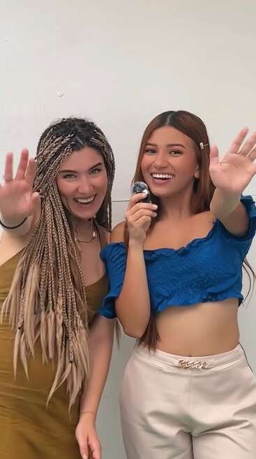 Deux jeunes femmes sourient joyeusement à la caméra. L'une a de longues tresses et porte une robe marron ; l'autre porte un haut bleu et tient un micro. Ambiance conviviale.