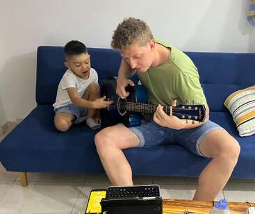 Un homme et un jeune garçon sont assis sur un canapé bleu. L'homme joue de la guitare tandis que le garçon montre du doigt avec enthousiasme, tous deux semblant absorbés et heureux dans leur activité.