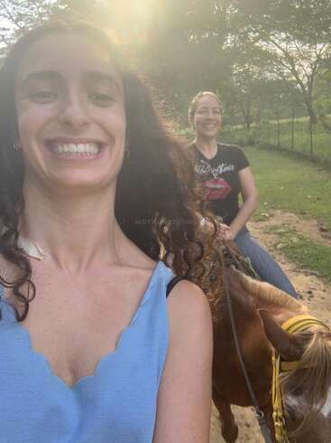 Deux femmes sourient joyeusement dans un décor ensoleillé en plein air. L'une prend un selfie, tandis que l'autre monte un cheval brun. Elles sont entourées d'herbe verte et d'arbres.