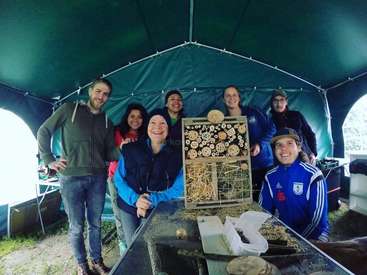 A imagem mostra um grupo de pessoas em pé ao redor de uma mesa com um hotel para insetos, sob uma tenda verde, sorrindo para a câmera.