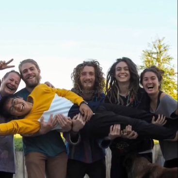 The image depicts a group of six people posing for a photo, with three individuals holding a fourth person aloft, set against a serene outdoor backdrop.
