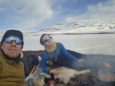 A man and woman relax outdoors in snowy mountains, wearing sunglasses and winter clothing, sitting on animal skins by a campfire with food, under a blue sky.
