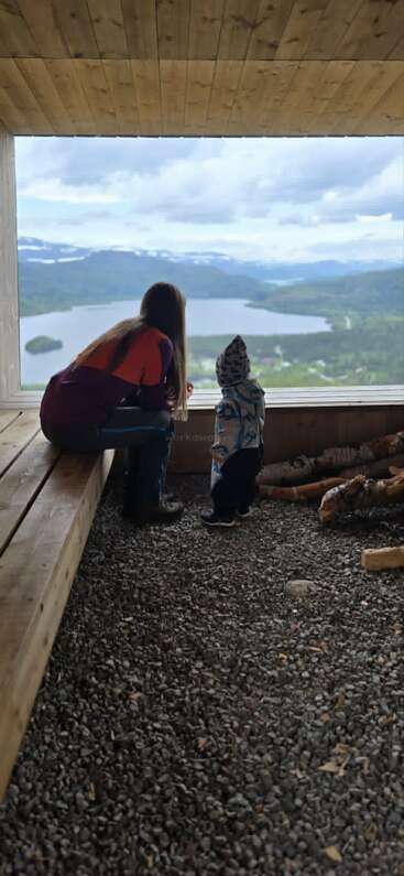 A woman and a small child sit in a wooden structure, gazing out at a scenic, mountainous lake landscape under a cloudy sky, sharing a quiet moment.