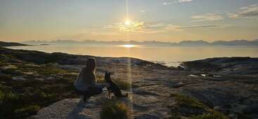 A woman crouches beside her dog, overlooking a serene lake and distant mountains at sunset. The sky reflects golden hues, creating a peaceful, tranquil scene.