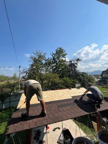 Two men are working on installing brown shingles on a wooden roof under a clear blue sky, surrounded by trees and greenery on a sunny day.
