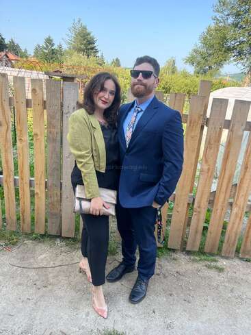 A couple poses together outdoors in front of a wooden fence, dressed formally. The woman holds a beige clutch, while the man wears a blue suit and sunglasses.