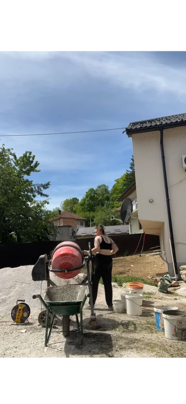 A woman stands outdoors operating a concrete mixer, filling a wheelbarrow with cement. Buckets and construction supplies are scattered nearby, with houses and greenery in the background.