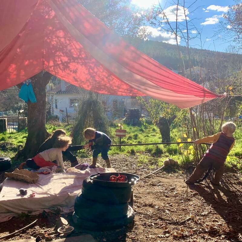 Três crianças brincam ao ar livre sob um grande toldo de tecido vermelho. A luz do sol penetra entre as árvores, projetando sombras. Elas parecem ter muita imaginação, curtindo a natureza e a companhia umas das outras.