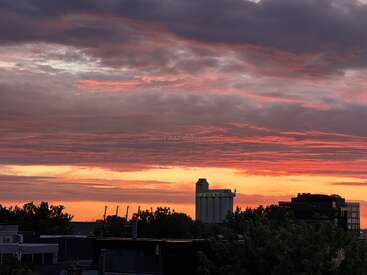 Un espectacular cielo al atardecer, con vibrantes tonos anaranjados, rojos y morados, se extiende sobre los edificios y los árboles de la ciudad, creando una silueta urbana tranquila pero impresionante.