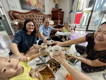 A happy family sits on the floor, sharing a meal and raising cups in a celebratory toast, with smiles, children, and delicious food in a cozy living room.