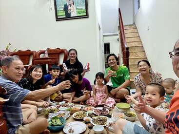 A joyful family gathers on the floor around a meal, smiling and sharing food, drinks, and laughter in a cozy, warmly lit home with stairs.