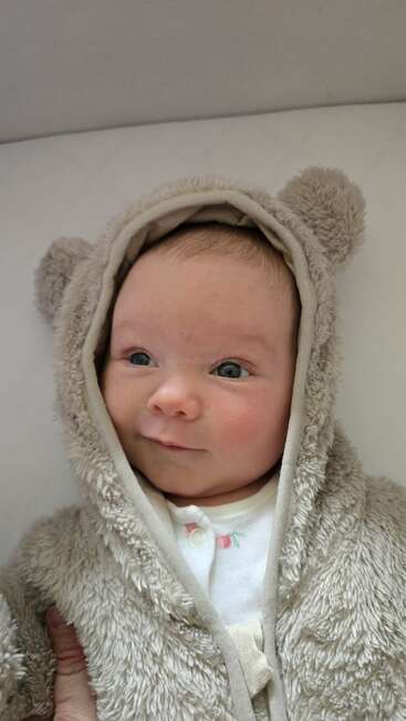 A cute baby is bundled in a soft, furry, light brown hooded outfit with bear ears, smiling gently while lying down, looking cozy and content.