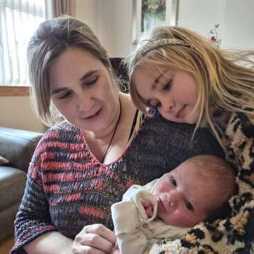 A woman lovingly holds a newborn baby while an older girl, possibly a sibling, gently leans in close, all sharing a tender family moment at home.