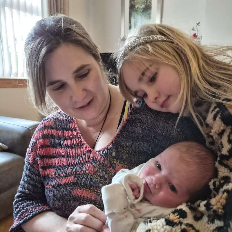 A woman lovingly holds a newborn baby while an older girl, possibly a sibling, gently leans in close, all sharing a tender family moment at home.