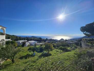 Um sol brilhante brilha sobre uma colina verde exuberante, com vista para casas brancas, árvores e mar azul cintilante sob um céu claro, criando uma vista cênica tranquila.