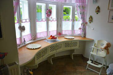 A cozy kitchen nook with white-framed windows, pink gingham curtains, decorative wreaths, a wooden countertop, fruit bowl, plates, high chair, and soft teddy bear.