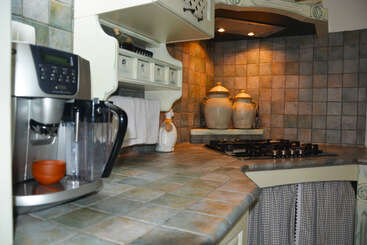 This image shows a cozy, tiled kitchen with a coffee machine, gas stove, decorative jars, under-shelf lighting, and fabric curtains covering the lower cabinets. Warm ambiance.