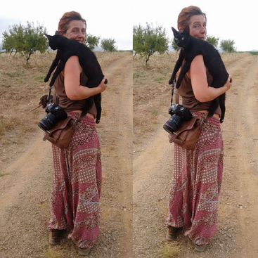 Eine Frau steht draußen auf einem Feldweg und hat eine schwarze Katze über der Schulter. Sie hat gemusterte Hosen an und trägt eine Kamera. Das Bild ist doppelt zu sehen.
