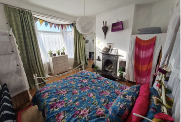 Helles, vielseitiges Schlafzimmer mit Erkerfenster, grünen Vorhängen, bunter Blumenbettdecke, Kamin und fröhlichen Deko-Elementen. Pflanzen und Sonnenlicht sorgen für eine warme, gemütliche und einladende Atmosphäre.