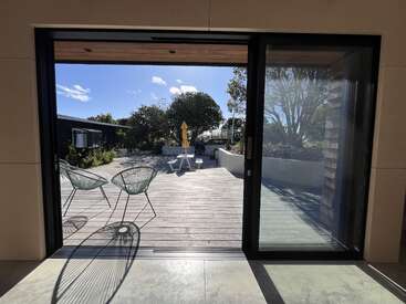 Dieses Bild zeigt eine sonnige Terrasse, die man durch große Glastüren sieht. Zwei moderne Stühle werfen Schatten. Bäume, ein Picknicktisch und ein gelber Sonnenschirm bilden den Hintergrund.